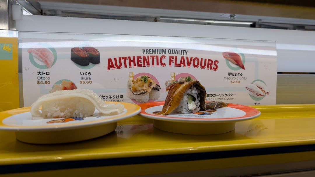 Fresh sushi dishes on a conveyor belt with a promotional display in the background.