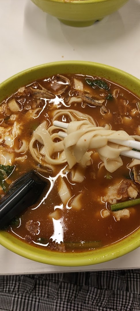 A close-up of a hearty bowl of noodles with rich broth, meat, and vegetables, held by chopsticks.