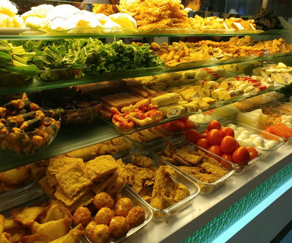 A vibrant display of fresh vegetables, fried snacks, and various ingredients in a multi-tiered food counter.