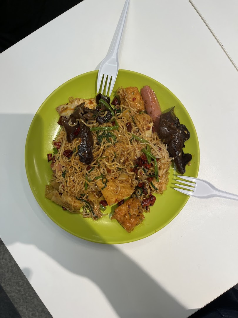 A vibrant plate of spicy noodles with various toppings, including mushrooms, tofu, and sausage, served with plastic forks.