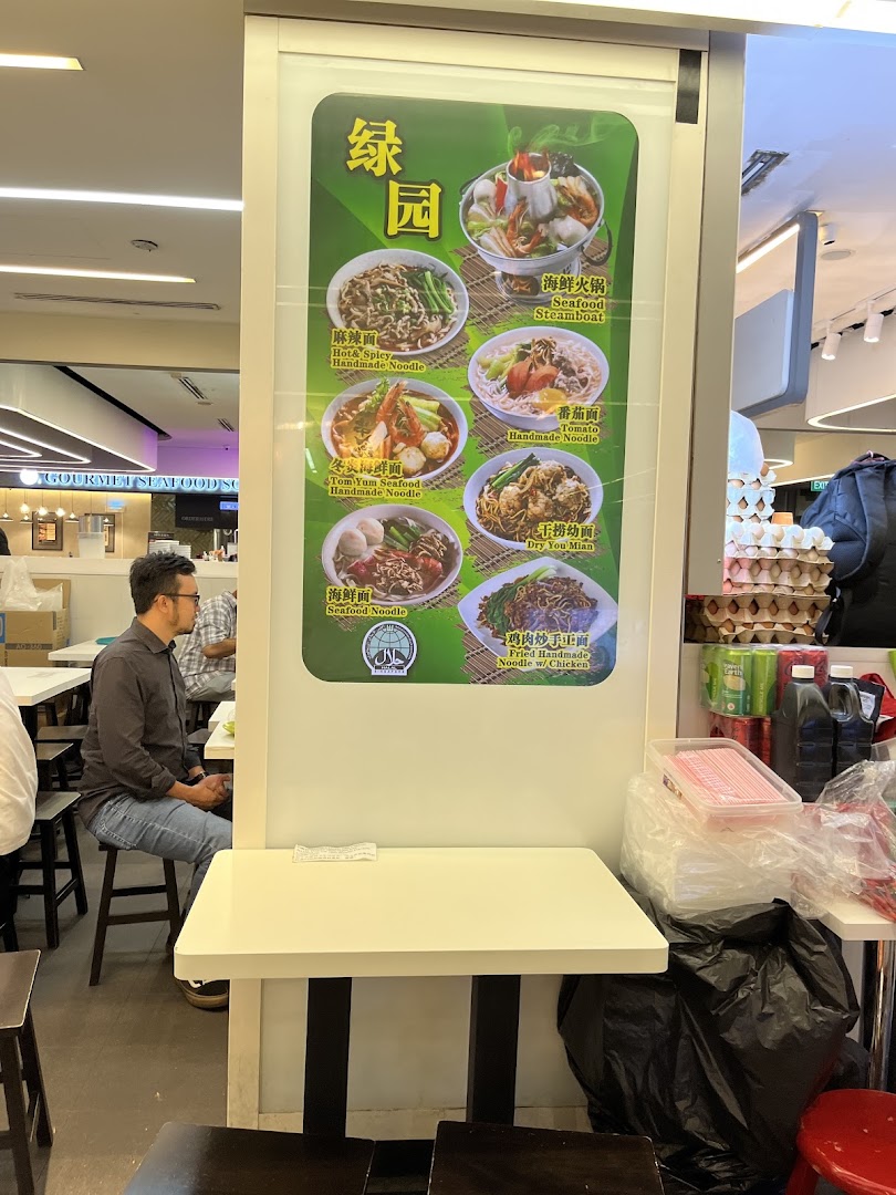 A large menu board at Green Delights showcasing various noodle dishes and a steamboat, with customers seated nearby.