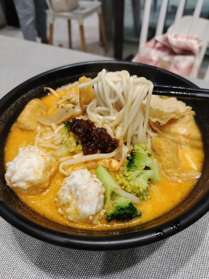 A close-up of a vibrant bowl of noodles with rich broth, fish balls, broccoli, and chili paste.