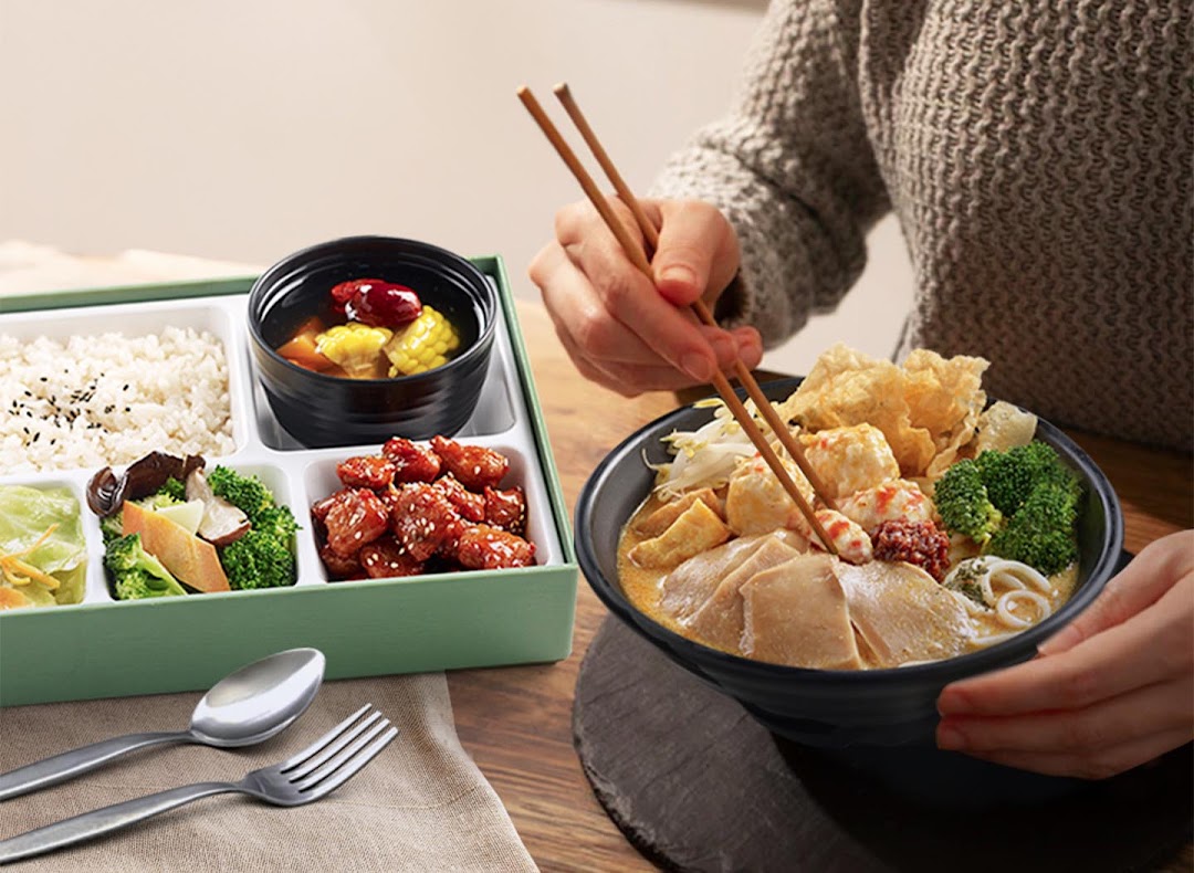 A person enjoying a vibrant bento box and a large bowl of noodle soup with chopsticks.