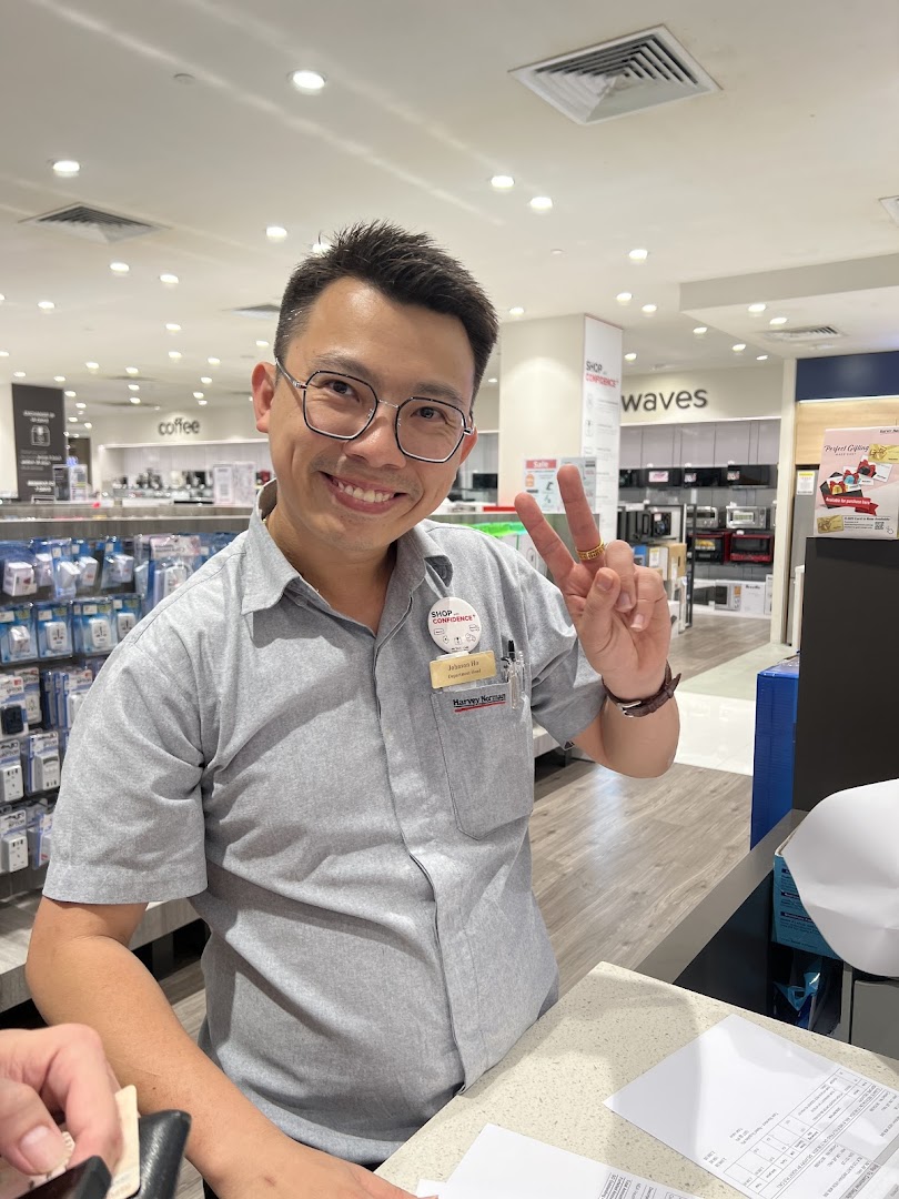 A friendly Harvey Norman staff member smiles and makes a peace sign inside the electronics and appliance store.