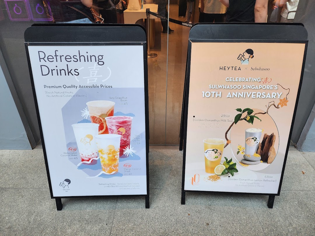 Two A-frame signs outside HEYTEA, showcasing refreshing drinks and a special collaboration menu.
