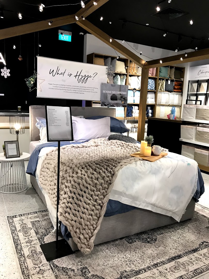 A cozy bed display featuring a chunky knit blanket, candles, and a 'What is Hygge?' sign in a home decor store.