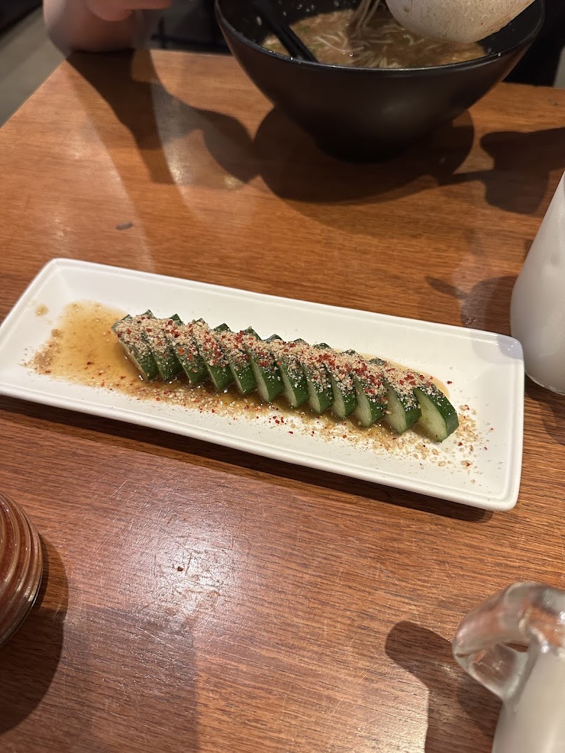 A plate of sliced cucumbers with sauce and toppings on a wooden table, with a bowl of ramen in the background.