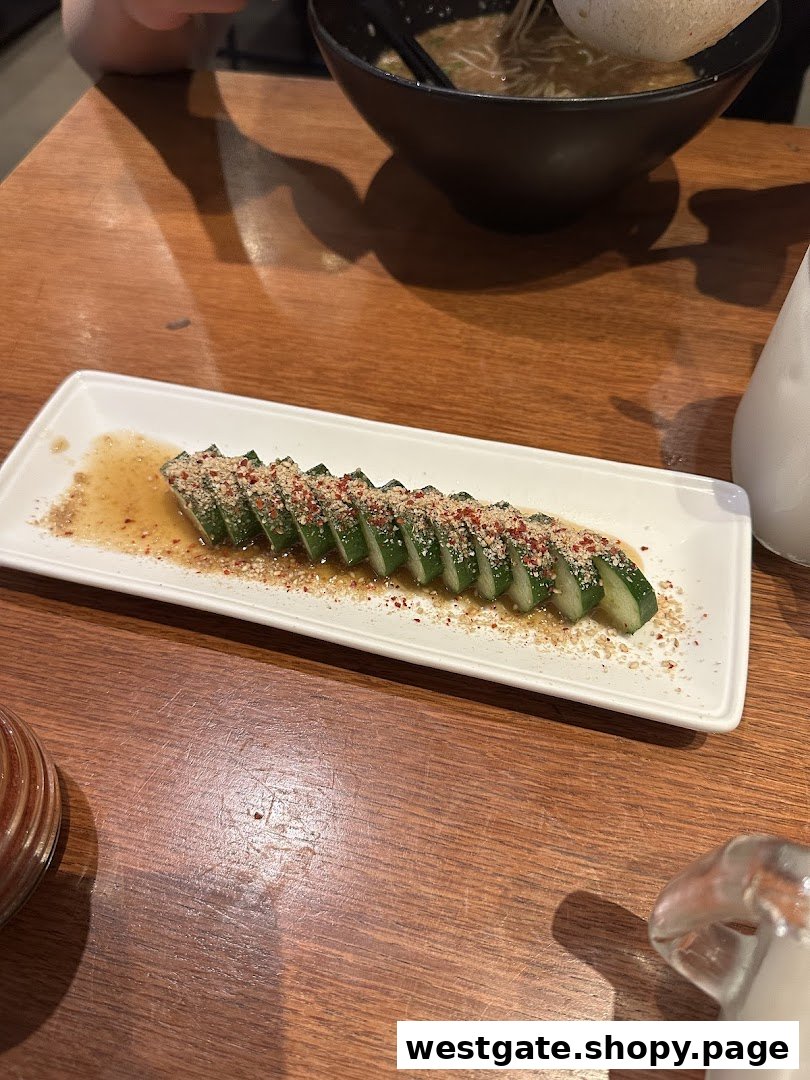 A plate of sliced cucumbers with sauce and toppings on a wooden table, with a bowl of ramen in the background.
