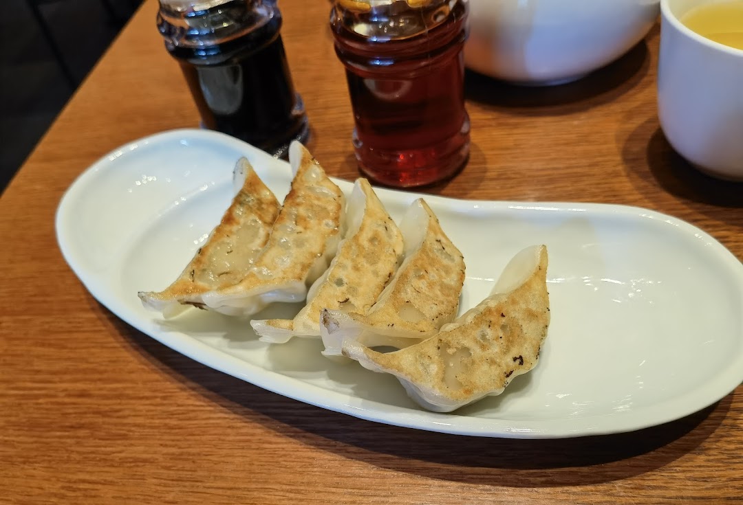 A close-up of perfectly pan-fried gyoza served on a white plate, with condiments and a drink in the background.