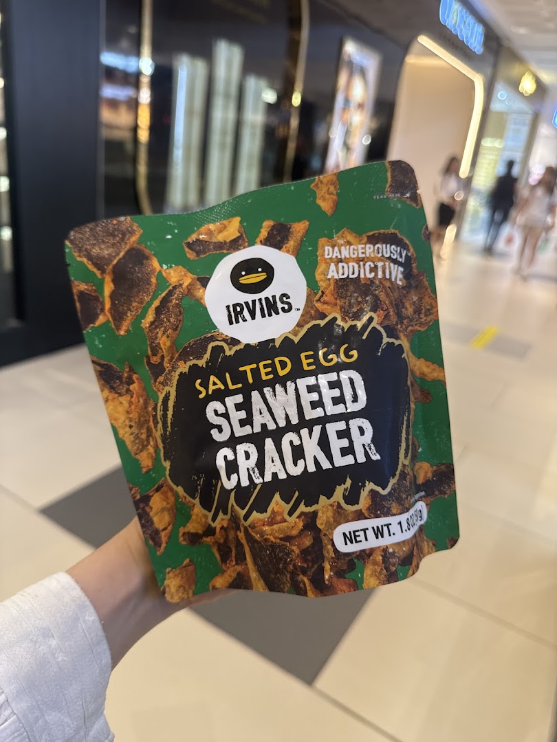 A hand holds a bag of IRVINS Salted Egg Seaweed Cracker in a mall setting.