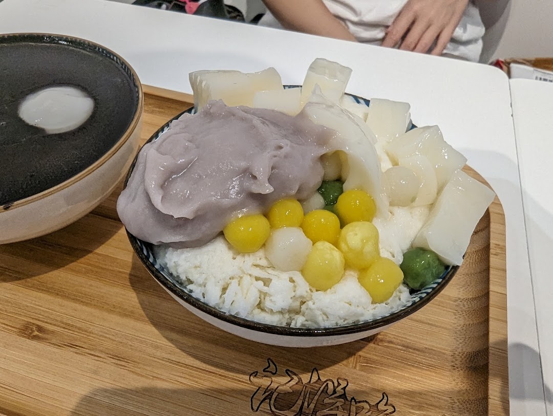 A vibrant bowl of shaved ice dessert with taro paste, various toppings, and a separate black sesame soup.