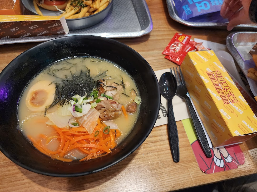 A close-up of a delicious ramen bowl with egg, chicken, and vegetables, alongside a JINJJA Corndog box.