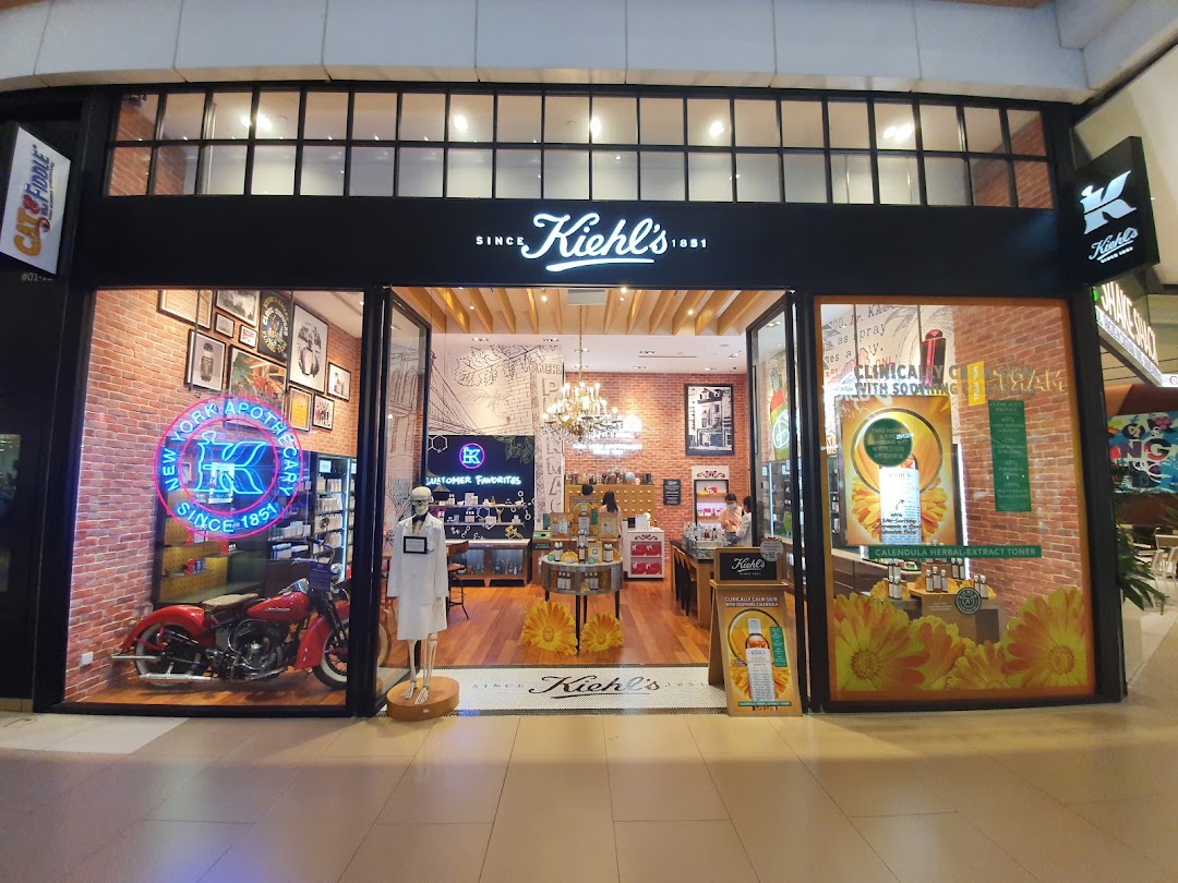 The storefront of Kiehl's Westgate, featuring a vintage motorcycle, neon sign, and product displays.