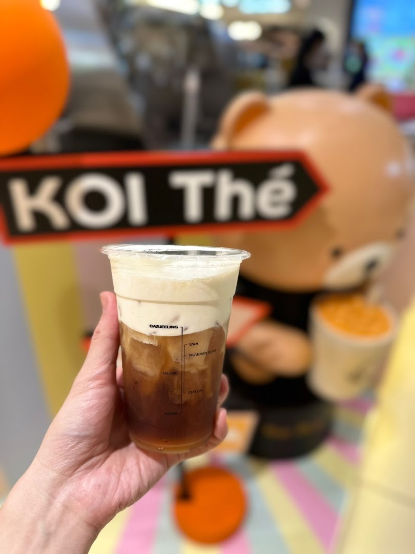 Hand holding a KOI Thé cream foam tea, with tea origins marked on the cup, in front of a shop display.