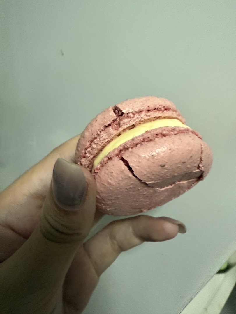A hand holds a pink macaron with a creamy yellow filling, showing its texture.