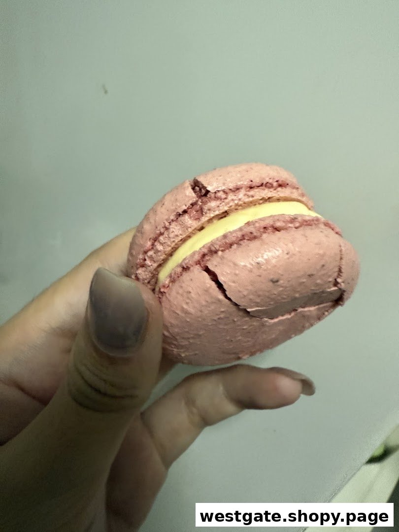 A hand holds a pink macaron with a creamy yellow filling, showing its texture.