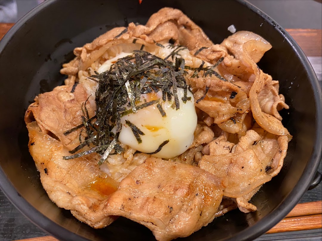 A close-up of a Japanese rice bowl featuring grilled pork, a soft-boiled egg, and shredded nori.