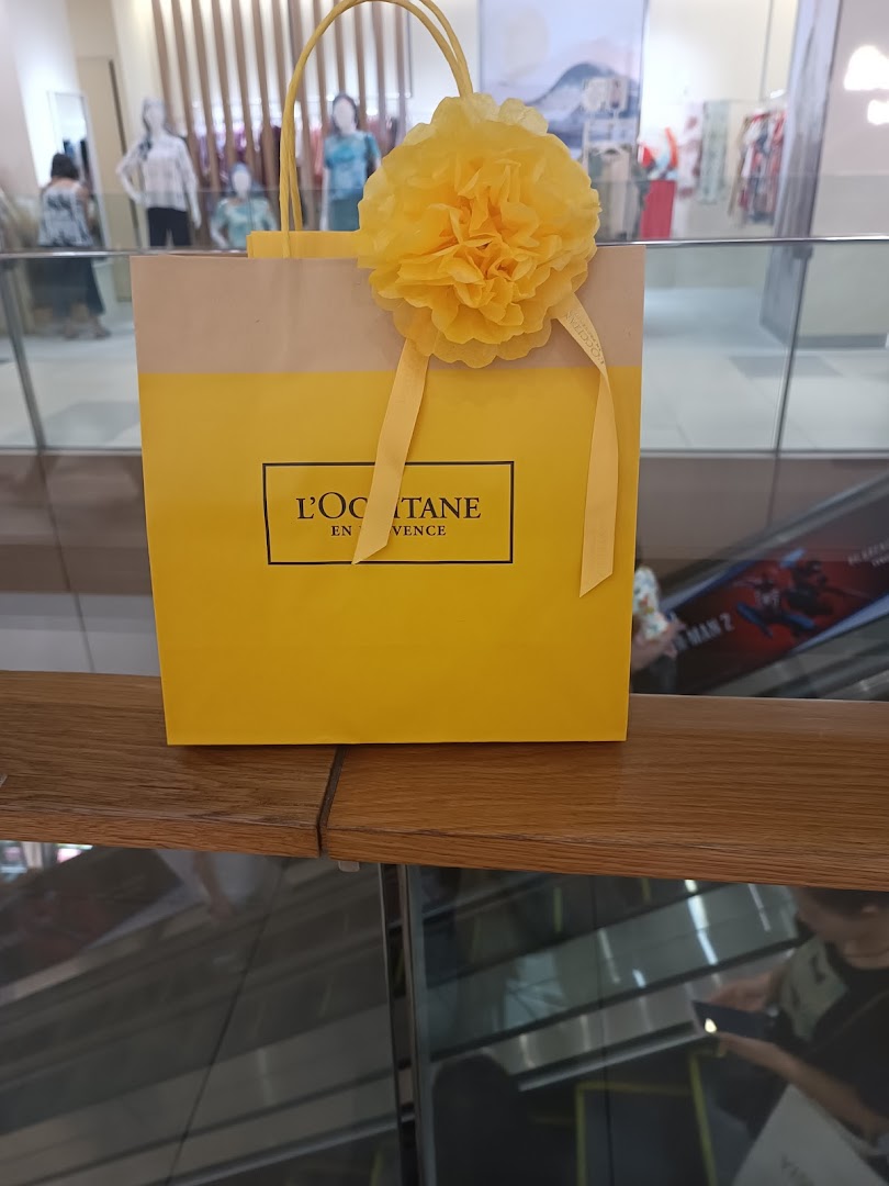 A bright yellow L'OCCITANE shopping bag adorned with a decorative yellow flower, set against a mall background.