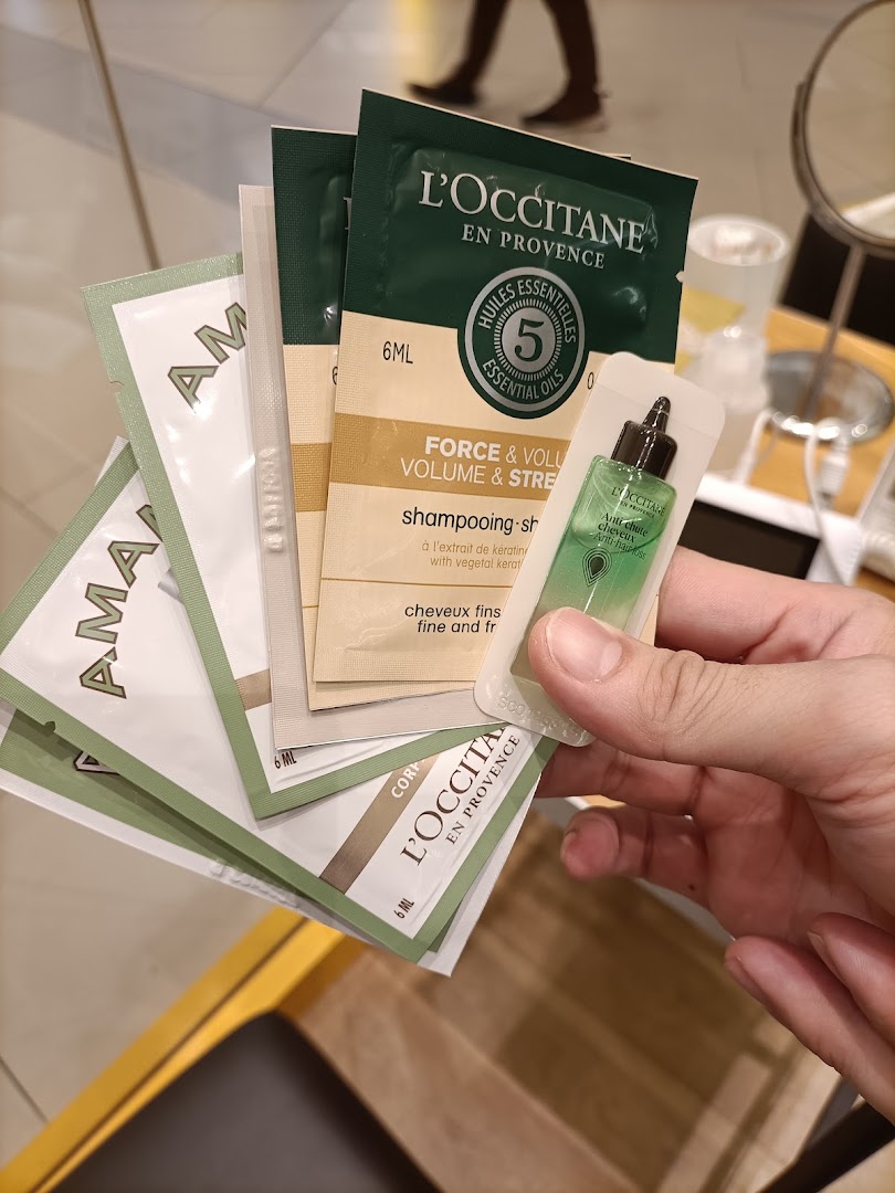 A hand holds various L'Occitane product samples, including shampoo and anti-hair loss treatments.