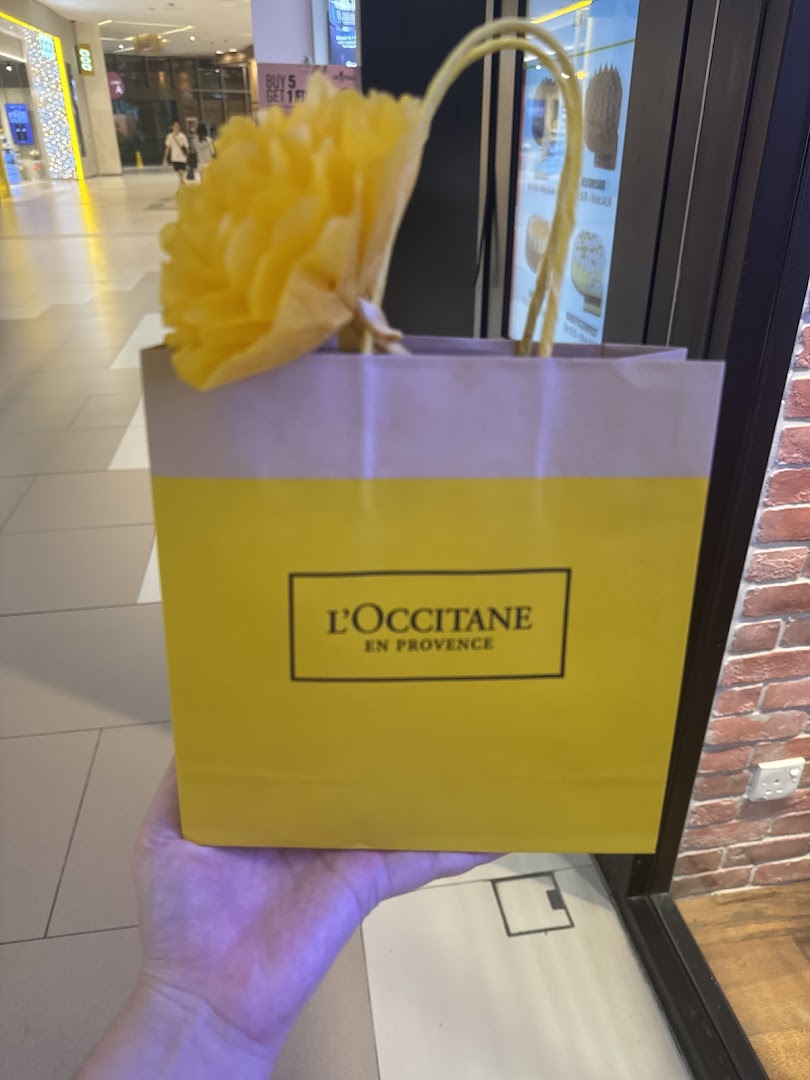 A person holds a vibrant yellow L'OCCITANE shopping bag adorned with a decorative flower inside a bustling mall.