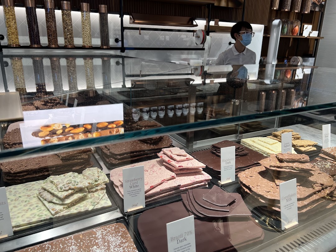 A vibrant display of fresh chocolate slabs and pralines in a glass case at Läderach, with a staff member visible.