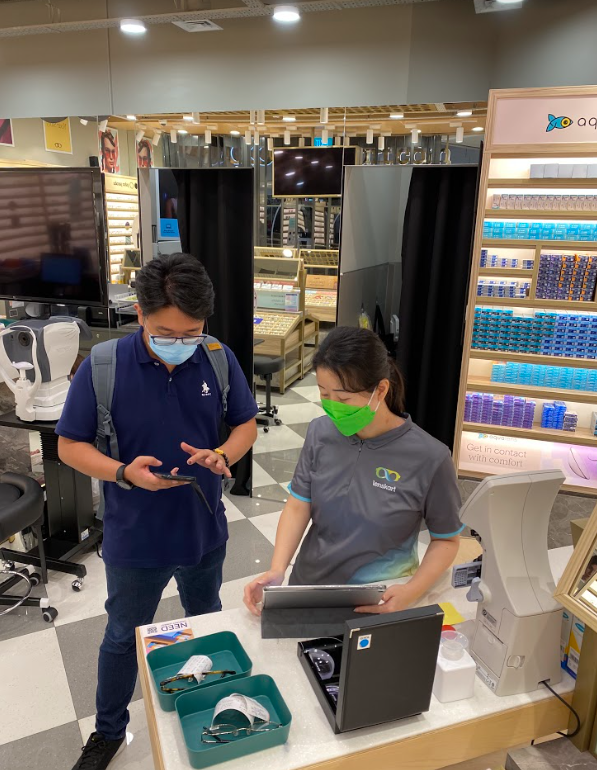 A customer consults with a Lenskart staff member inside the eyewear store, surrounded by optical equipment and products.