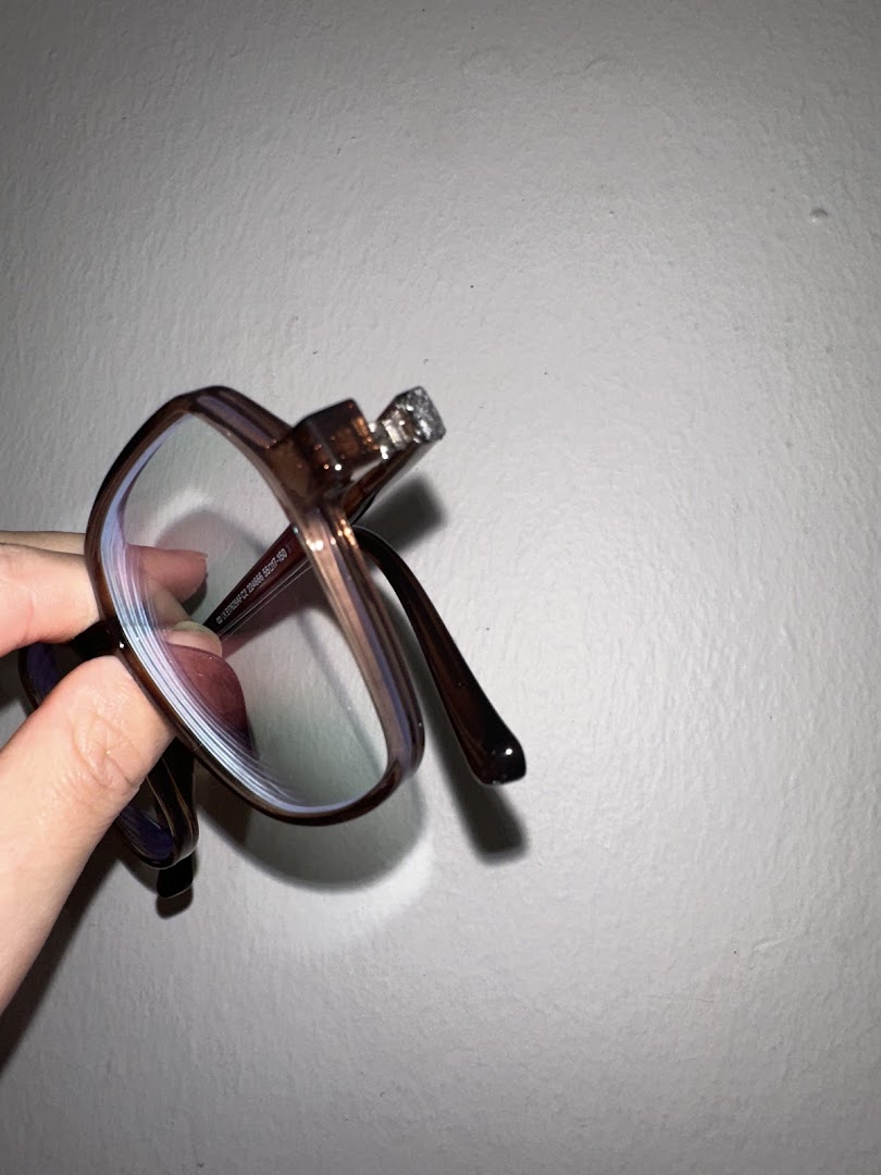 A hand holds a pair of stylish brown eyeglasses with clear lenses against a white wall.