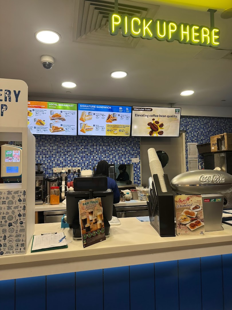 A bright service counter with digital menu boards, a 'PICK UP HERE' sign, and a staff member.
