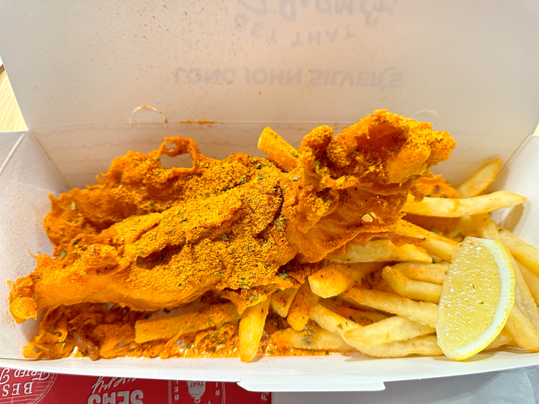 A close-up of crispy, seasoned fried fish and golden fries with a lemon wedge in a takeaway box.
