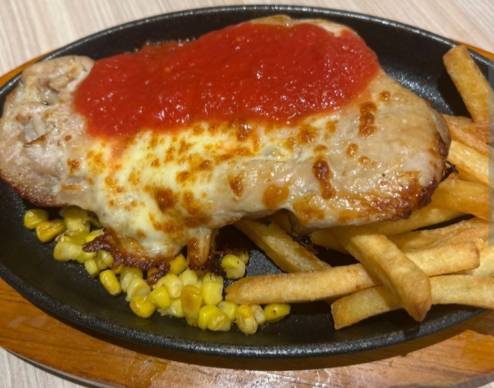 A sizzling plate featuring a cheesy chicken cutlet topped with tomato sauce, served with fries and corn.