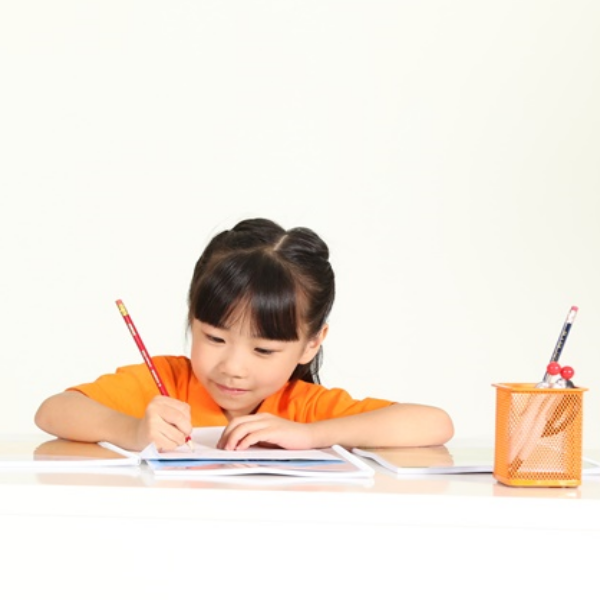 A young girl in an orange shirt diligently writing at a desk with a pencil.