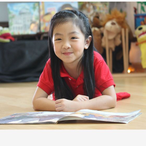 A young girl in a red shirt smiles while reading a book on the floor.