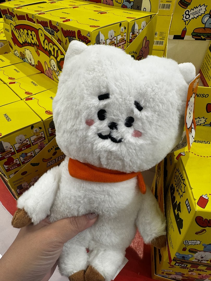 A hand holds a white fluffy BT21 RJ plush toy in MINISO, surrounded by yellow 'Burger Party' themed product boxes.