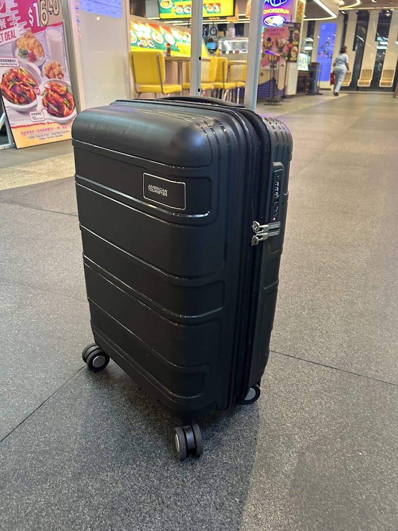 A black American Tourister suitcase stands on a tiled floor within a bustling shopping mall.