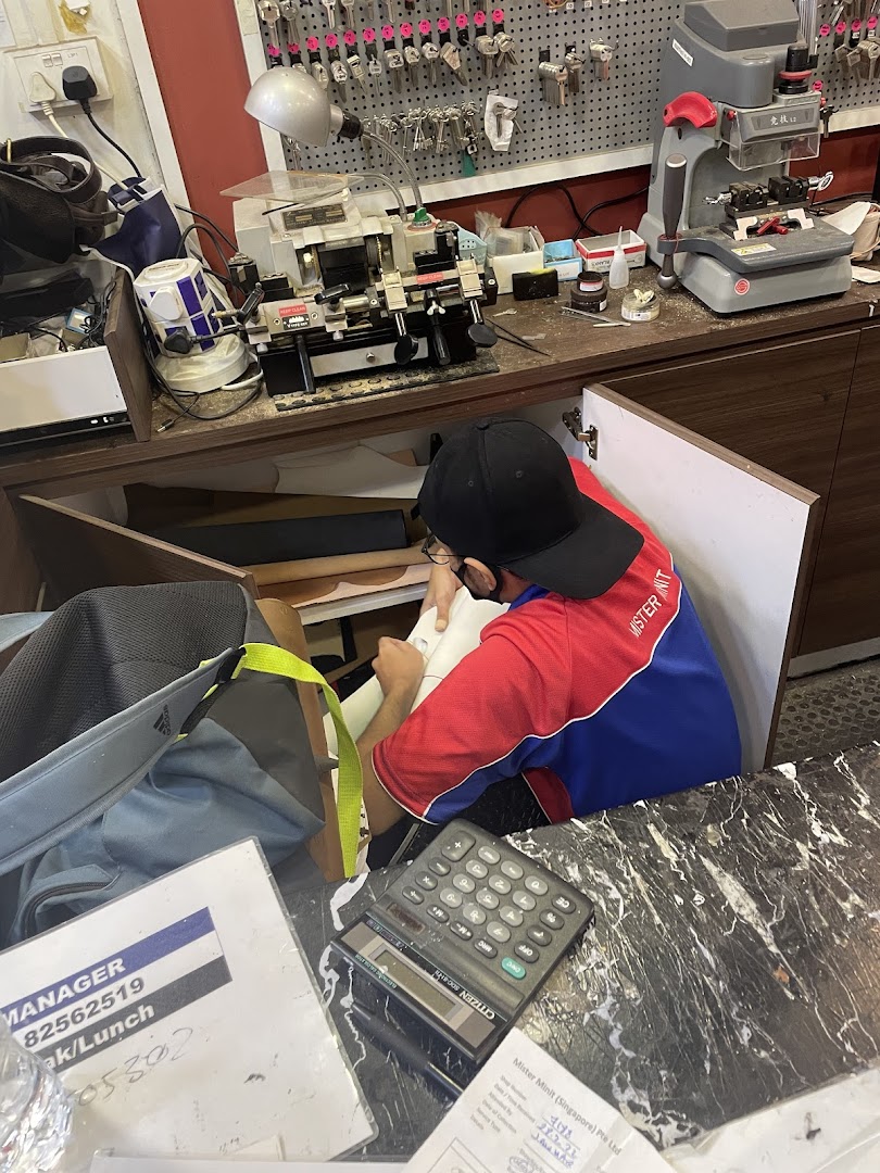 A Mister Minit staff member works at a counter with key cutting and repair equipment.