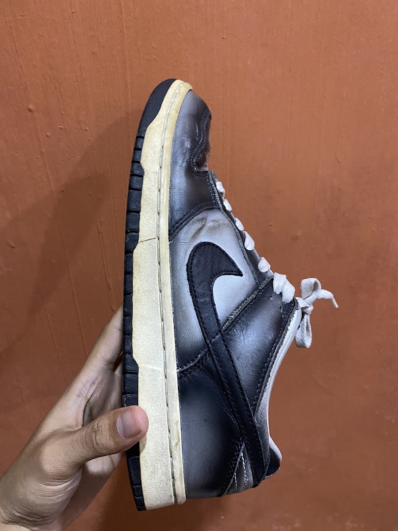 A hand holds a worn black and grey sneaker, showcasing its side profile against an orange background.