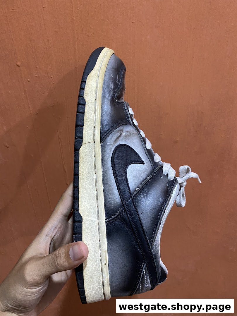 A hand holds a worn black and grey sneaker, showcasing its side profile against an orange background.