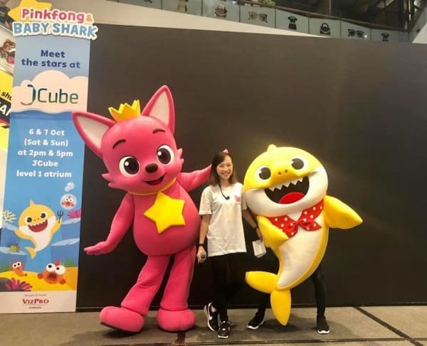 A woman poses with Pinkfong and Baby Shark mascots next to an event banner at JCube.