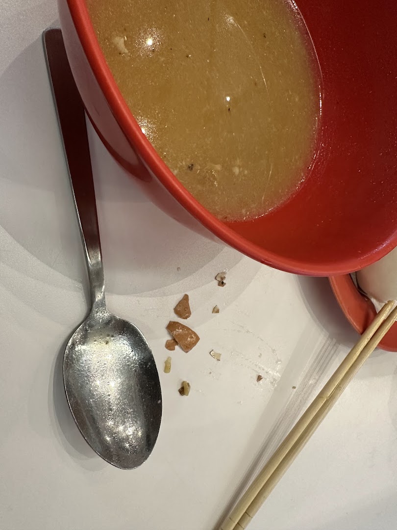 A red bowl of soup with a spoon and chopsticks on a white table, showing food remnants.