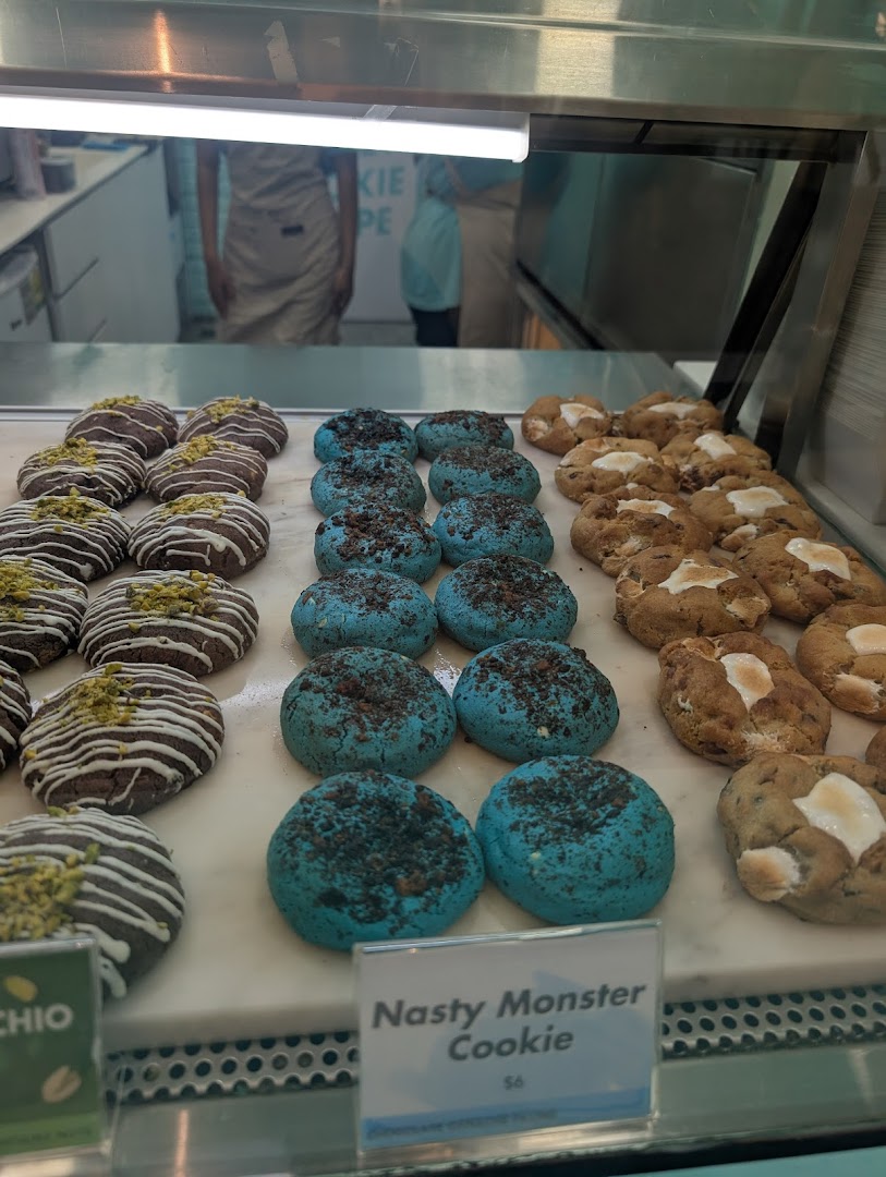 A vibrant display of gourmet cookies, including blue 'Nasty Monster Cookies' and chocolate pistachio varieties, in a bakery showcase.