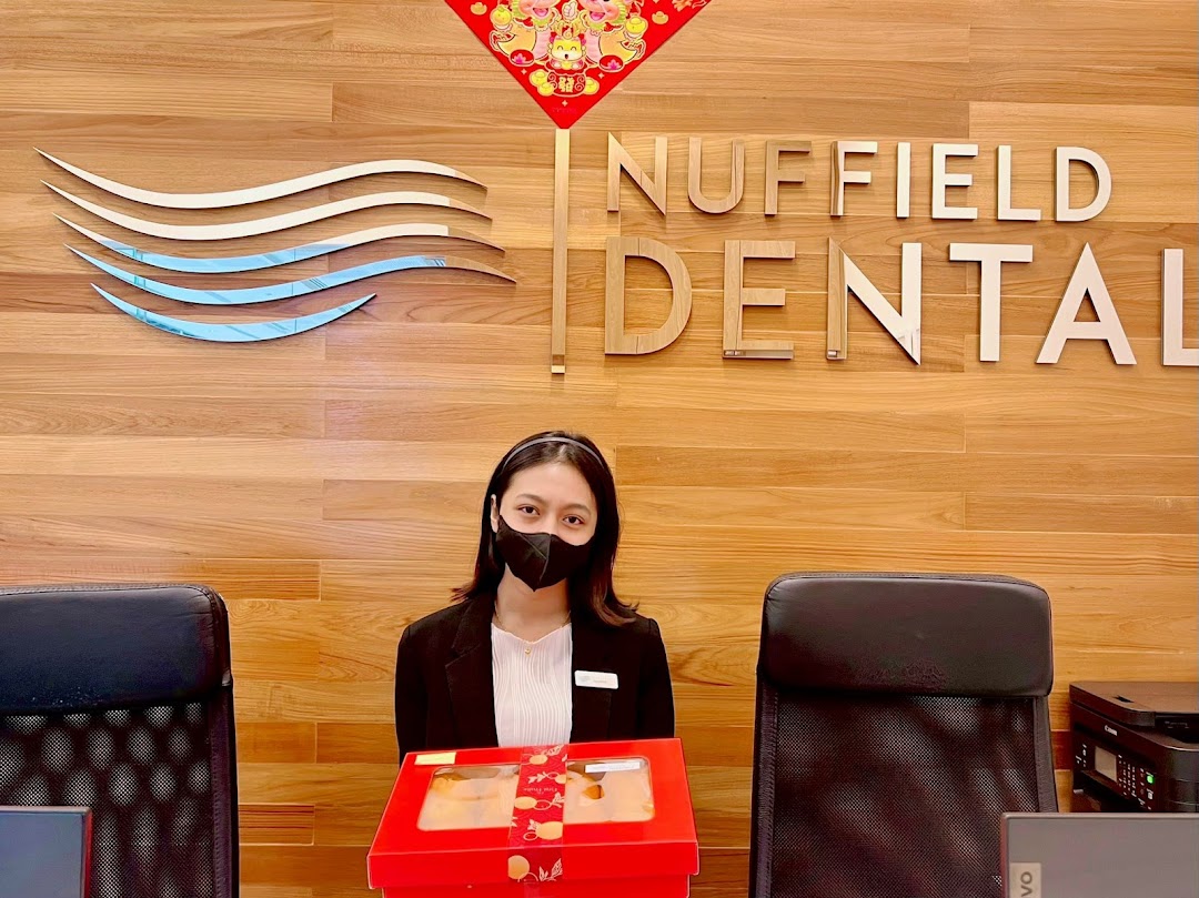 A masked staff member at the Nuffield Dental reception desk with the clinic's logo and name.