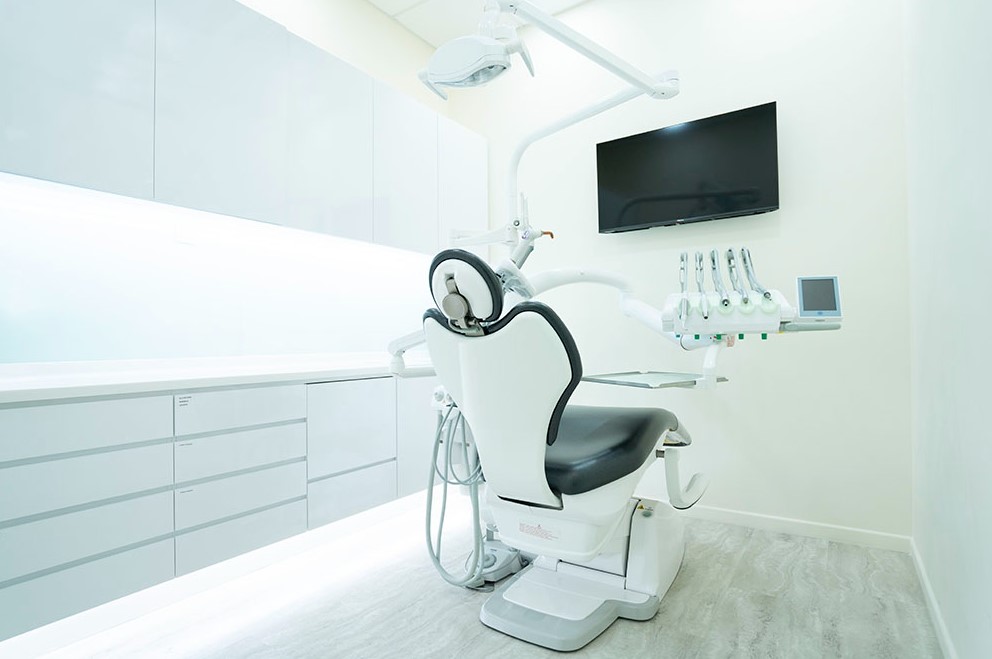 A modern, clean dental treatment room with a patient chair, overhead light, and dental tools.