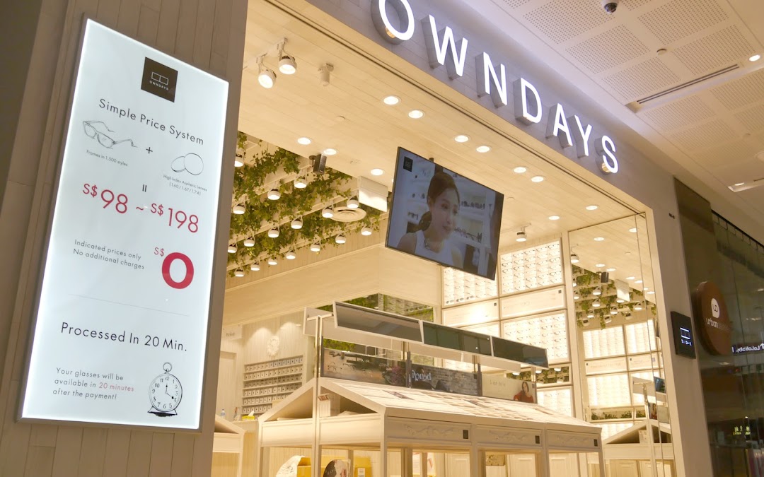 A bright OWNDAYS shop entrance featuring a prominent sign detailing their simple eyewear pricing system and quick processing.