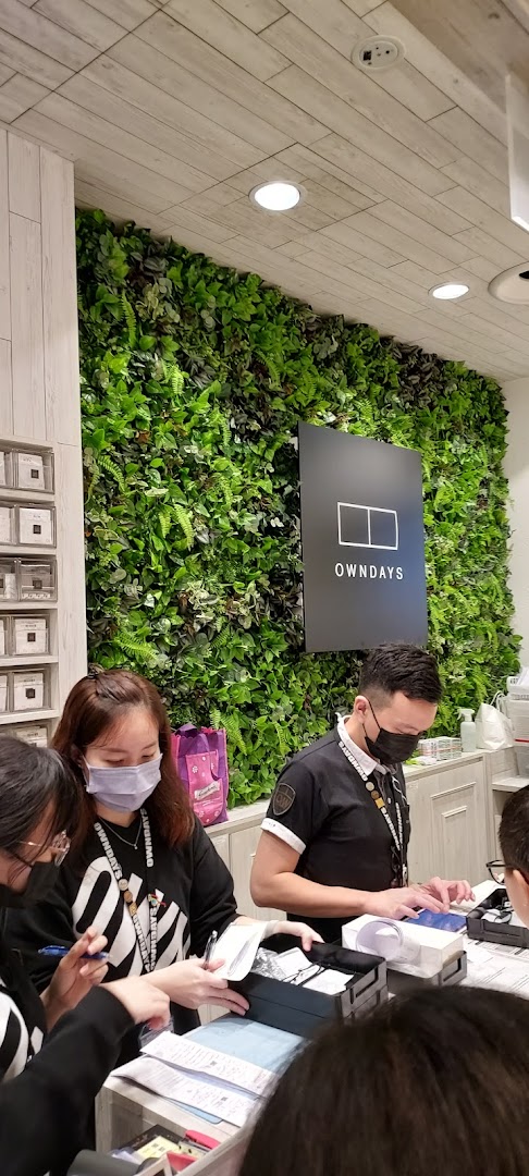 Customers and staff interacting at the counter of OWNDAYS, featuring a prominent green wall and brand logo.
