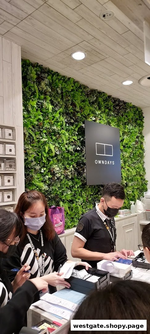Customers and staff interacting at the counter of OWNDAYS, featuring a prominent green wall and brand logo.