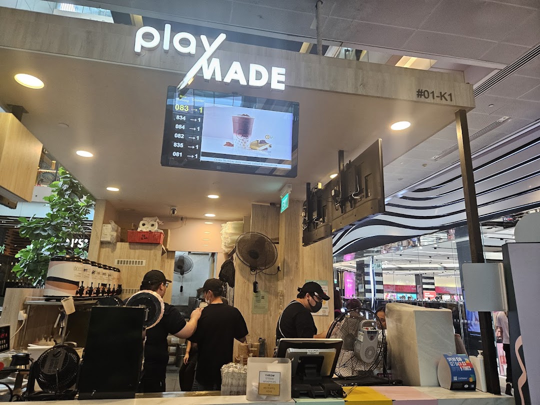 An interior view of PlayMade bubble tea shop, showing staff preparing drinks and a digital order display.