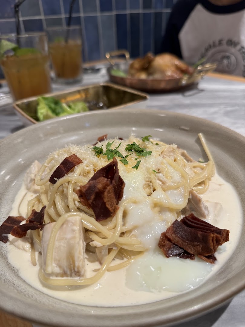 A close-up of creamy pasta with chicken, bacon, and a poached egg, with drinks and roasted chicken in the background.
