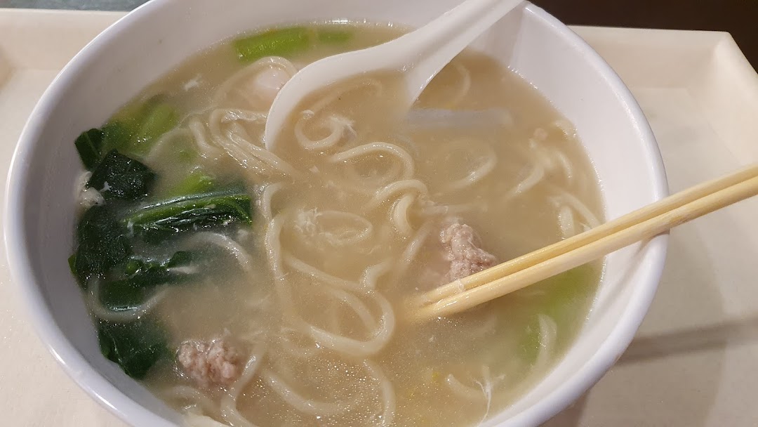 A close-up of a steaming bowl of noodles with broth, vegetables, and minced meat, served with chopsticks and a spoon.