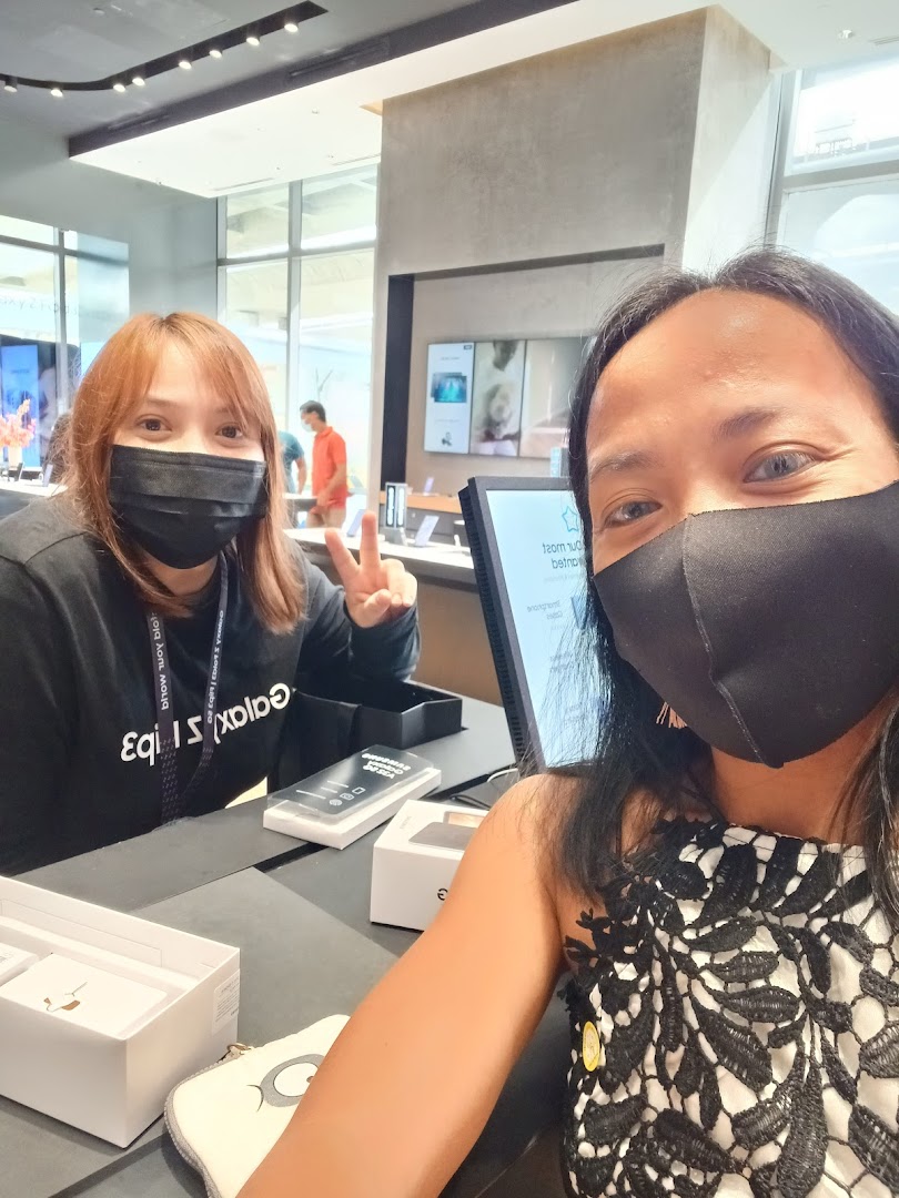 Two masked women, one staff and one customer, at a counter in a Samsung Experience Store, with products visible.
