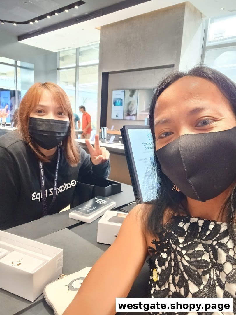 Two masked women, one staff and one customer, at a counter in a Samsung Experience Store, with products visible.